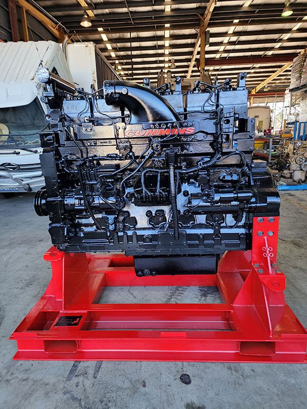 Rebuilt Cummins diesel engine, freshly painted and bolted to a red workshop stand, with a truck parked behind it in the shed