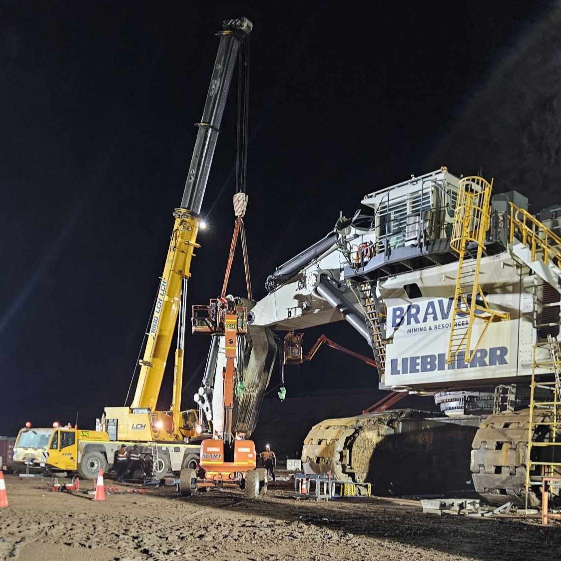A Liebherr mining excavator under repair at night, a mobile crane lifting part of its digging arm while crews work from elevated work platforms