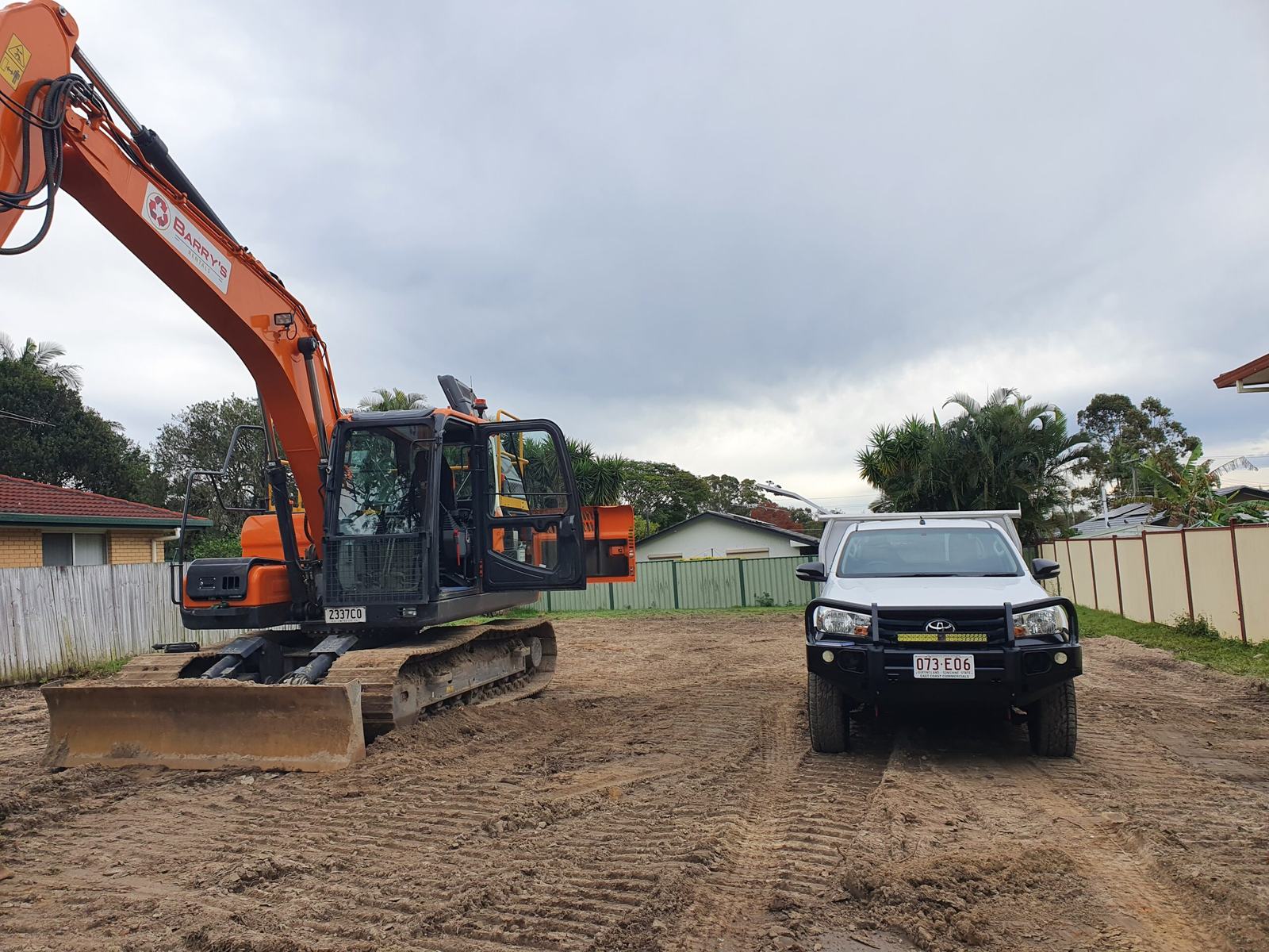 A white service ute parked beside an orange tracked excavator on a cleared dirt block