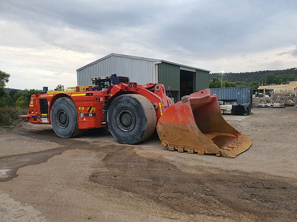 An orange underground mining loader with a large front bucket, parked on a gravel yard in front of a steel shed